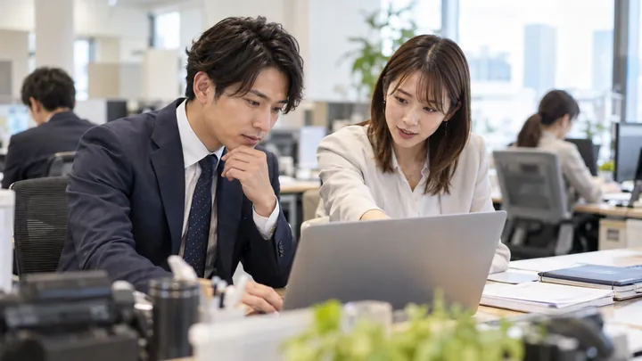 営業担当がノートパソコンで提案資料を確かめ、隣の同僚が画面を指さして助言している